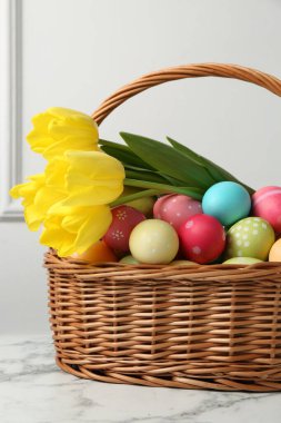 Hasır sepete boyanmış Paskalya yumurtaları ve beyaz duvarın yanındaki beyaz mermer masada güzel laleler.