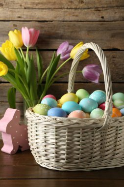 Hasır sepete boyanmış Paskalya yumurtaları, dekoratif tavşan figürü ve ahşap masada güzel laleler.