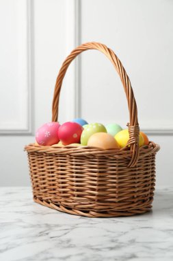 Hasır sepete boyanmış Paskalya yumurtaları beyaz duvarın yanındaki beyaz mermer masada.