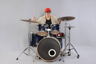 Gri arka planda davul çalmayı öğrenen, gözlüklü ve şapkalı gülümseyen genç çocuk.