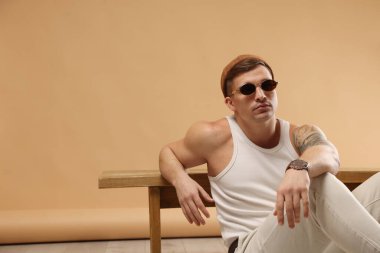 Moda modeli. Şapkalı ve güneş gözlüklü yakışıklı. Yerde poz veriyor. Bankta bej arka plana yaslanıyor.