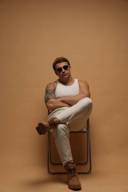 Moda modeli. Şapkalı ve güneş gözlüklü yakışıklı, bej arka planda çapraz kollu sandalyede poz veriyor.