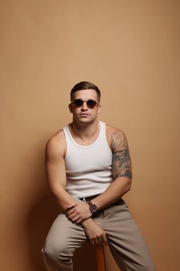 Moda modeli. Güneş gözlüklü yakışıklı adam bej arka planda taburede poz veriyor.