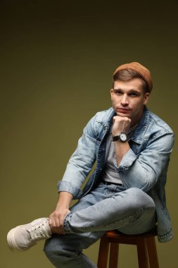 Moda modeli. Şapkalı ve ceketli yakışıklı adam, koyu yeşil arka planda dışkı üzerinde poz veriyor.