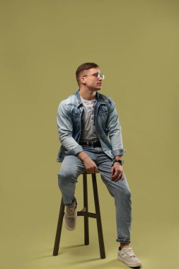 Moda modeli. Yeşil arka planda taburede poz veren gözlüklü ve ceketli yakışıklı adam.