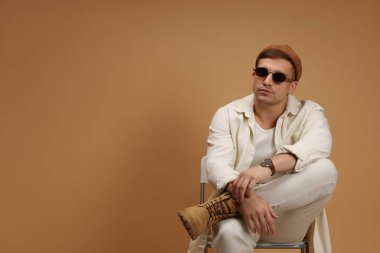 Moda modeli. Şapkalı ve güneş gözlüklü yakışıklı adam bej arka plana karşı sandalyede poz veriyor, mesaj için yer var.