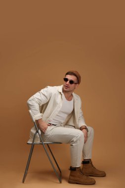 Moda modeli. Şapkalı ve güneş gözlüklü yakışıklı bej arka planda sandalyede poz veriyor.