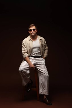 Moda modeli. Siyah arka planda taburede poz veren güneş gözlüklü yakışıklı bir adam.