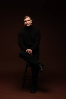 Moda modeli. Koyu kahverengi arka planda taburede poz veren yakışıklı bir adam.