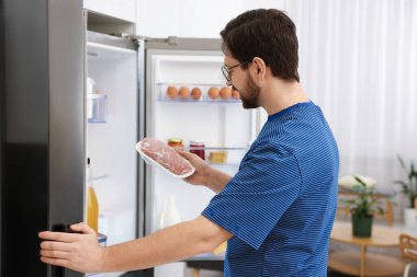 Mutfaktaki açık buzdolabının yanında çiğ et yiyen adam.