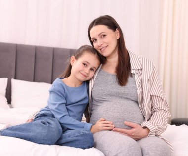 Hamile kadın ve kızı evde yatakta oturuyor.