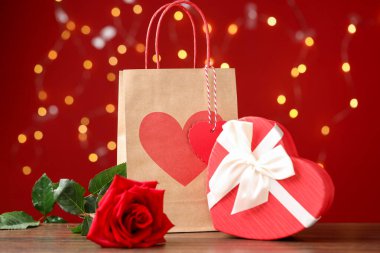 Kağıt torba, gül ve kalp şeklinde hediye paketi ahşap masada kırmızı arka planda bulanık ışıklarla