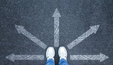 Spor ayakkabılı bir adam yolda duruyor, oklar farklı yönlere bakıyor, üst manzara. Yolculuğuna başla.