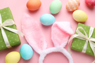 Boyalı Paskalya yumurtaları, tavşan kulaklıkları ve pembe arka planda hediyeler.