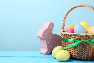 Hasır sepete boyanmış Paskalya yumurtaları ve açık mavi arka planda ahşap masa üzerindeki dekoratif tavşan figürü. Metin için boşluk