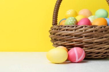Hasır sepetin içine boyanmış Paskalya yumurtaları beyaz masada, sarı arka planda, yakın plan. Metin için boşluk