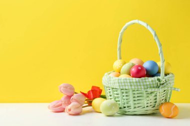 Hasır sepete boyanmış Paskalya yumurtaları ve sarı arka planda beyaz masada güzel laleler. Metin için boşluk