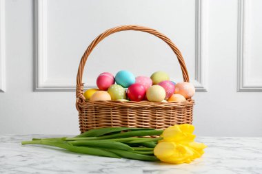 Hasır sepete boyanmış Paskalya yumurtaları ve beyaz duvarın yanındaki beyaz mermer masada güzel laleler.
