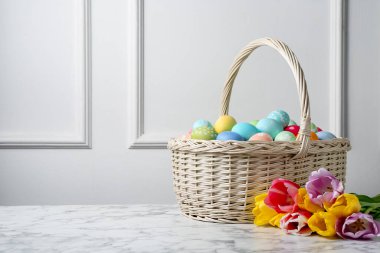 Hasır sepete boyanmış Paskalya yumurtaları ve beyaz duvarın yanındaki beyaz mermer masada güzel laleler. Metin için boşluk