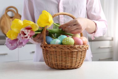 Paskalya sepetinde yumurta boyalı bir kadın ve beyaz masa içinde laleler, yakın plan.