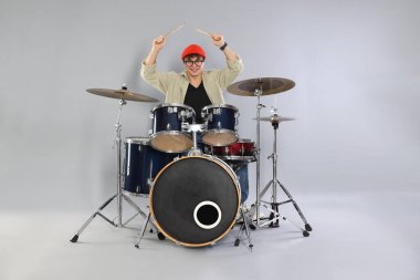 Gri arka planda davul çalmayı öğrenen, gözlüklü ve şapkalı gülümseyen genç çocuk.