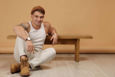 Moda modeli. Şapkalı yakışıklı adam bej arka plana yaslanarak yerde poz veriyor.