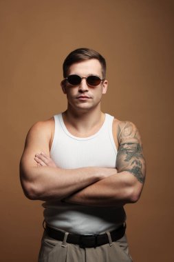 Moda modeli. Güneş gözlüklü yakışıklı bej arka planda çapraz kollarla poz veriyor.