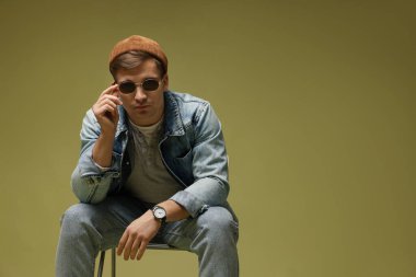 Moda modeli. Şapkalı, güneş gözlüklü ve ceketli yakışıklı adam koyu yeşil eğimli arka plana karşı sandalyede poz veriyor.