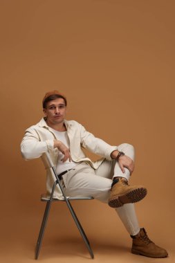 Moda modeli. Sandalyede bej arka planda poz veren şapkalı yakışıklı bir adam.