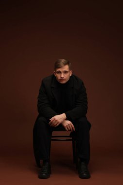 Moda modeli. Koyu kahverengi arka planda sandalyede poz veren yakışıklı bir adam.