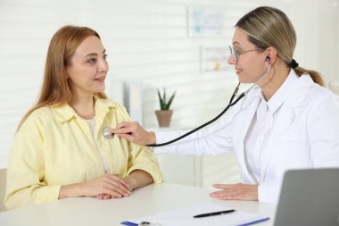 Steteskopla gülümseyen kardiyolog klinikteki hastanın kalp atışlarını dinliyor.