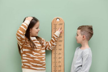 Küçük bir çocuk ve kız açık yeşil duvarın yanında boylarını grafikle ölçüyorlar.