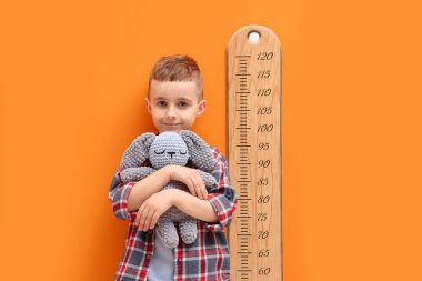 Oyuncak tavşancığı olan küçük çocuk turuncu duvarın yanında boyunu ölçüyor.