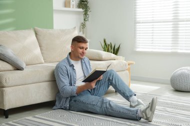 Evdeki kanepenin yanında kitap okuyan genç bir adam.