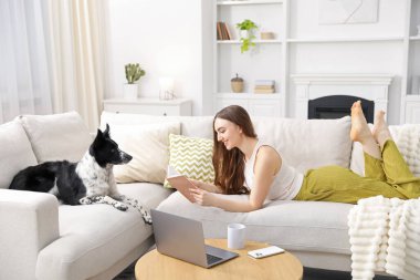 Sevimli köpeğiyle evdeki kanepede kitap okuyan gülümseyen kadın.