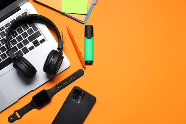 Kulaklık, dizüstü bilgisayar, akıllı telefon, turuncu arka planda akıllı saat ve kırtasiye malzemesi, düz yatak. Metin için boşluk