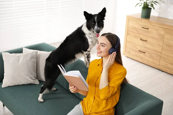 Kulaklıklı gülümseyen kadın kitap okuyor ve evdeki kanepede sevimli köpeği oturuyor.