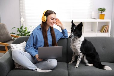 Kulaklıklı, dizüstü bilgisayarlı, sevimli köpeği evdeki kanepede gülümseyen kadın.