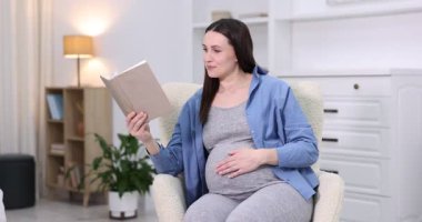 Hamile kadın evdeki koltukta kitap okuyor.