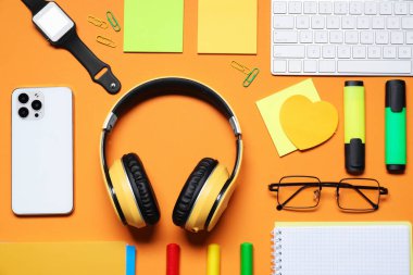 Kulaklıklar, bilgisayar klavyesi, akıllı telefon, akıllı saat, turuncu arka planda gözlük ve kırtasiye malzemesi, düz konum