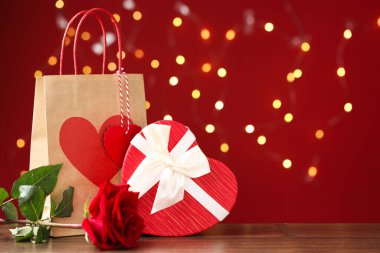 Kağıt torba, gül ve kalp şeklinde hediye paketi ahşap masada kırmızı arka planda bulanık ışıklarla. Metin için boşluk