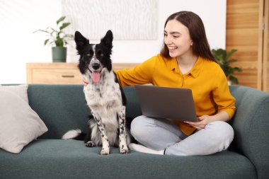 Evdeki kanepede dizüstü bilgisayarı ve sevimli köpeğiyle gülümseyen kadın.