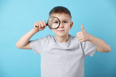 Büyüteci olan küçük çocuk açık mavi arka planda baş parmağını gösteriyor.