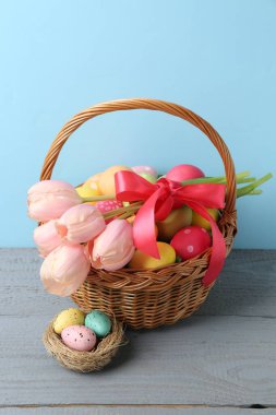 Hasır sepete boyanmış Paskalya yumurtaları ve açık mavi arka plana karşı gri ahşap masada güzel laleler.