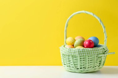 Hasır sepetin içine boyanmış Paskalya yumurtaları beyaz masada, sarı arka planda. Metin için boşluk