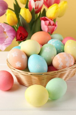 Hasır sepetin içine boyanmış Paskalya yumurtaları beyaz ahşap masa üzerinde ve sarı arka planda güzel laleler, yakın plan.
