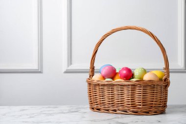 Hasır sepete boyanmış Paskalya yumurtaları beyaz duvarın yanındaki beyaz mermer masada. Metin için boşluk