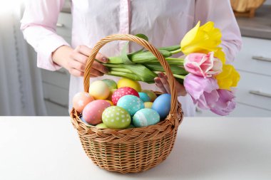 Paskalya sepetinde yumurta boyalı bir kadın ve beyaz masa içinde laleler, yakın plan.