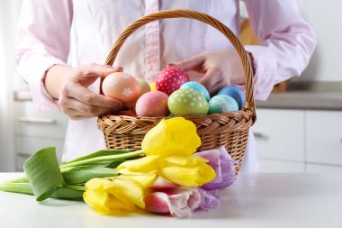 Paskalya sepetinde yumurta boyalı bir kadın ve beyaz masa içinde laleler, yakın plan.