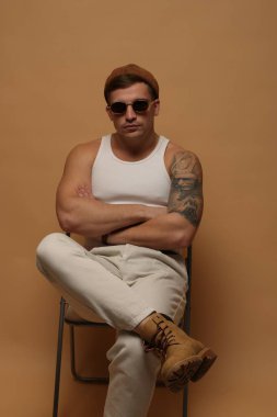 Moda modeli. Şapkalı ve güneş gözlüklü yakışıklı, bej arka planda çapraz kollu sandalyede poz veriyor.
